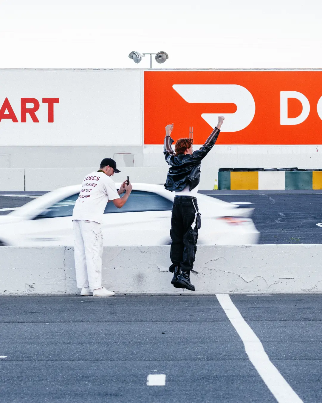 Zwei Personen machen ein Foto am Streckenrand, während ein weißer Hyundai Elantra N schnell an ihnen vorbeifährt. Die eine Person jubelt und springt dabei in die Luft.