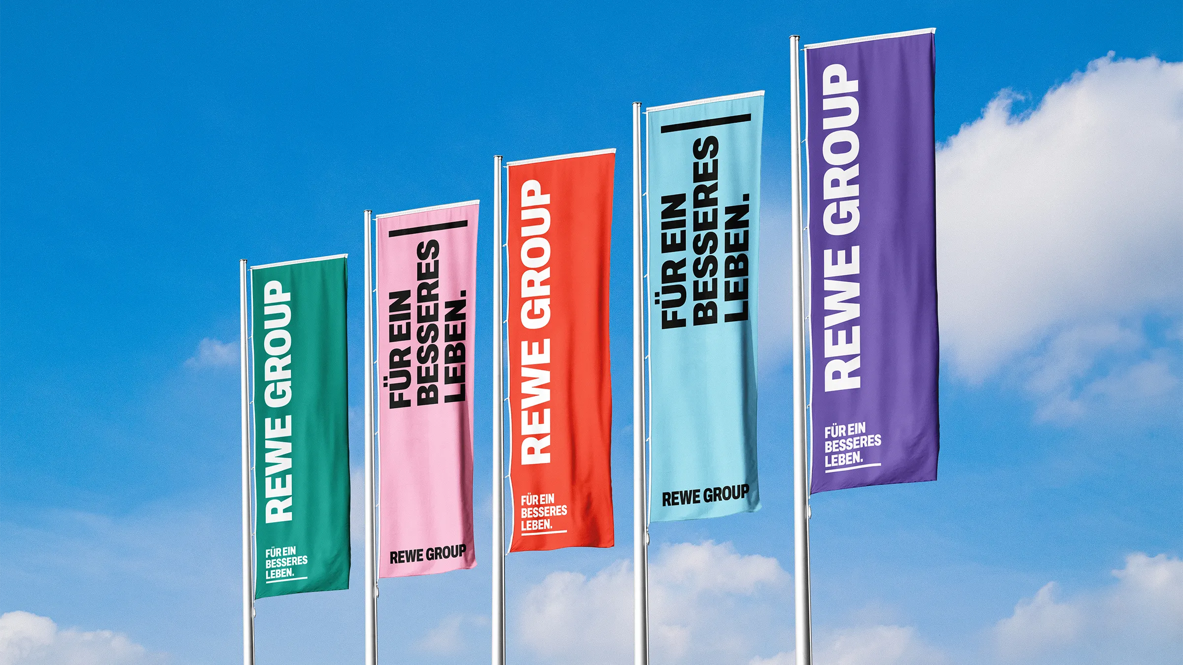Fünf Hochformat-Flaggen im neuen REWE GROUP Corporate Design vor blauem Himmel.